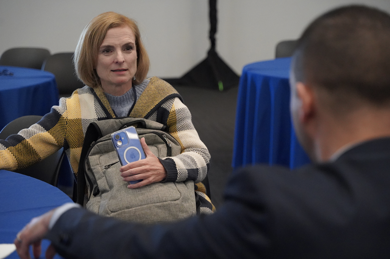A female attendee of a networking event sits with her phone and backpack, speaking to a male attendee whose back is turned to the viewer. There are royal blue standing table cloths behind them.